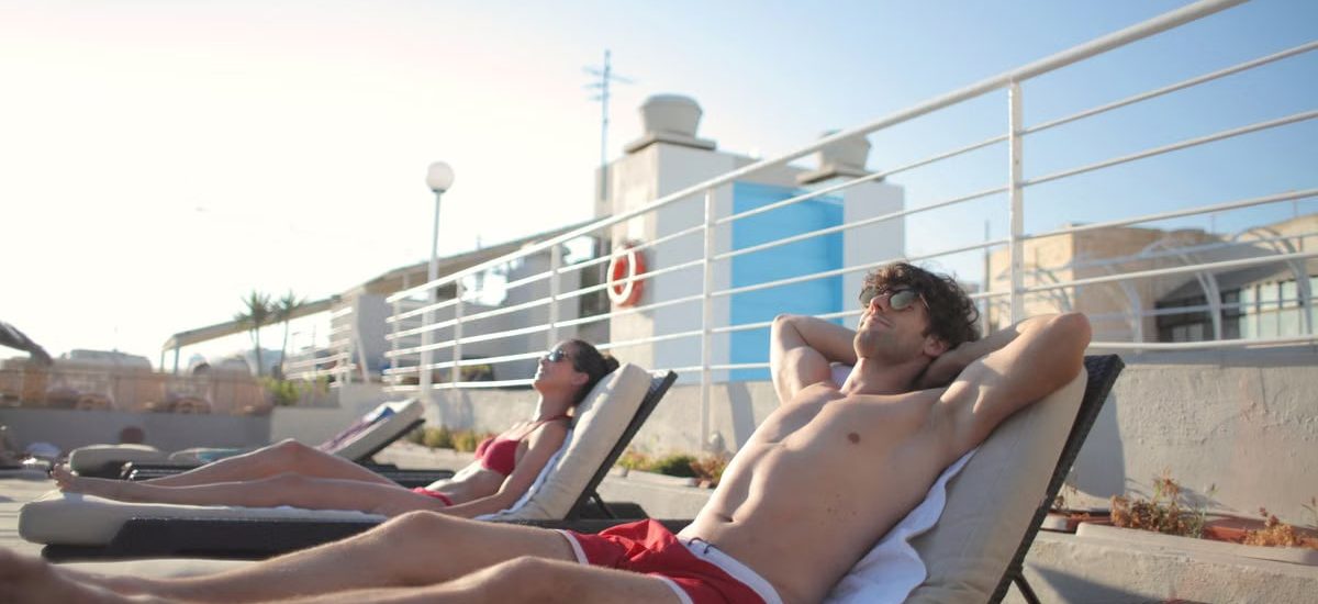 Shirtless male in red swimwear and sunglasses lying on sun loungers with hands under head next to sunbathing girlfriend in bikini on hot summer day